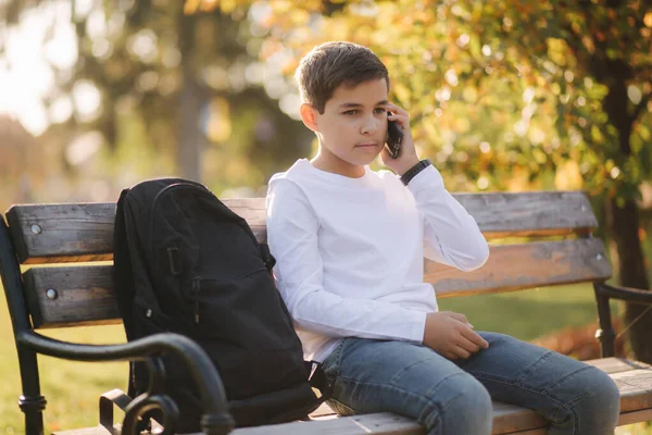 身穿白色运动衫 背着黑色背包的小男孩坐在公园的长椅上 与电话旁的某个人交谈 — 图库照片