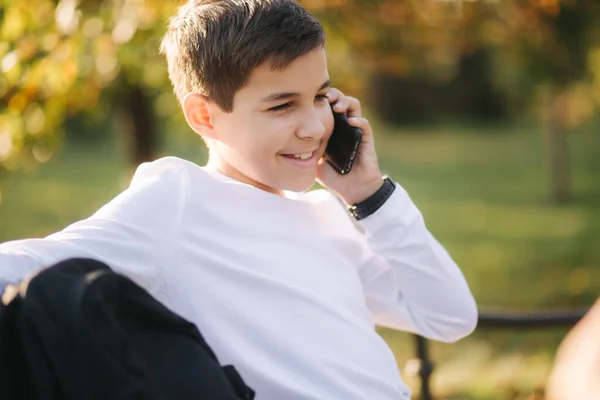 公園のベンチに座って電話で誰かと話す黒のバックパックと白いトレーナーの若い男の子 — ストック写真