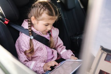 Beautiful little girl in car seat using digital tablet during trip. Happy female kid on back seat. High quality photo clipart