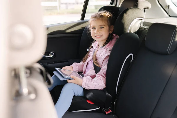 stock image Adorable child girl playing game with digital tablet while sitting in car seat of car during trip. Happy female kid enjoys vacation with tablet in car. High quality photo