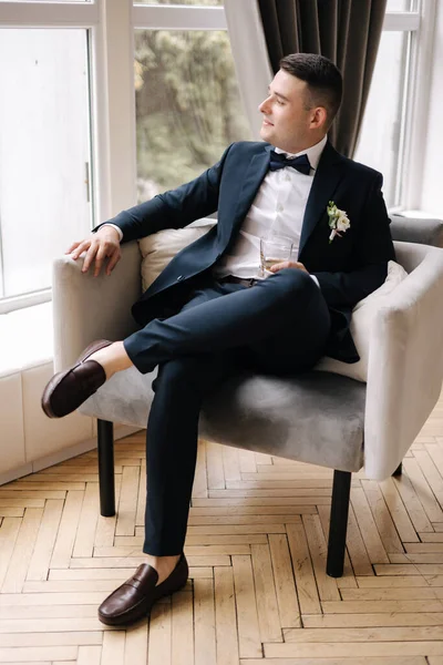 スタイリッシュな新郎の椅子に大きな窓の前に座っている 人はウイスキーを一杯飲む 結婚式の日 高品質の写真 — ストック写真
