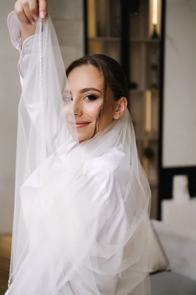 stock image Beautiful bride with long bridal veil dressed in grooms shirt stand in front of big window at home. High quality photo