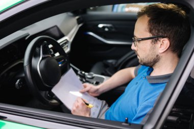 Araba teftişi sırasında erkek tamirci dokümanı doldururken yan görünüm. Adam arabanın içinde oturuyor. Yüksek kalite fotoğraf