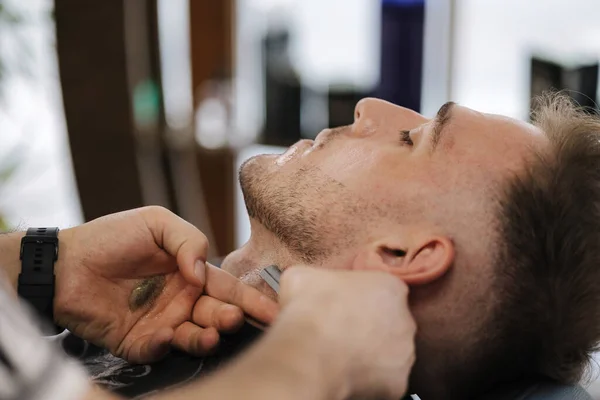 Barber using razor to shave beard for customer. Confident guy sitting at beauty salon. High quality photo