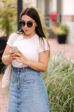 Kot etekli ve beyaz tişörtlü telefon kullanan güzel bir kadın. Kadın dışarıda dolaşıyor ve sosyal ağlarda bir şeyler arıyor. Yüksek kalite fotoğraf