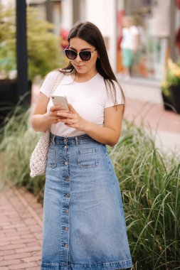 Kot etekli ve beyaz tişörtlü telefon kullanan güzel bir kadın. Kadın dışarıda dolaşıyor ve sosyal ağlarda bir şeyler arıyor. Yüksek kalite fotoğraf