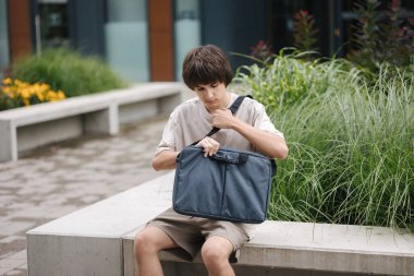 Genç çocuk bankta oturur ve laptop çantasını açar. Yüksek kalite fotoğraf