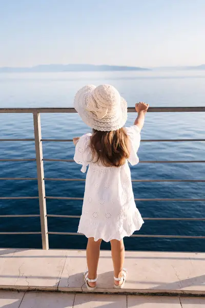 Adorable five year old girl in beautiful white dress and hat looking at the sea from height . High quality photo