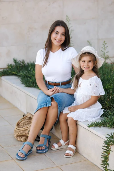 Mom and daughter sits on stone bench and smile. Beautiful little girl in white dress and hat. Mom in denim style. High quality photo
