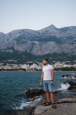 Yakışıklı sakallı adam uçurumun kenarında durur ve deniz batımını seyreder. Büyük dağların arkası. Yüksek kalite fotoğraf