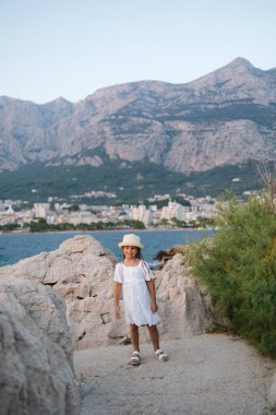 Deniz kıyısında fotoğrafçıya poz veren şirin küçük bir kız. Beyaz elbiseli ve örgü şapkalı güzel bir kız. Yüksek kalite fotoğraf