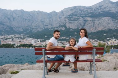 Mutlu aile portresi deniz kıyısında bankta oturur ve kameraya bakar. Deniz ve dağların arkası. Yüksek kalite fotoğraf