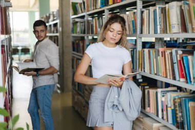 Yakışıklı çocuk kütüphanede güzel bir kıza bakıyor. Üniversite kütüphanesinde iki kişi. Kitap rafları. Yüksek kalite fotoğraf