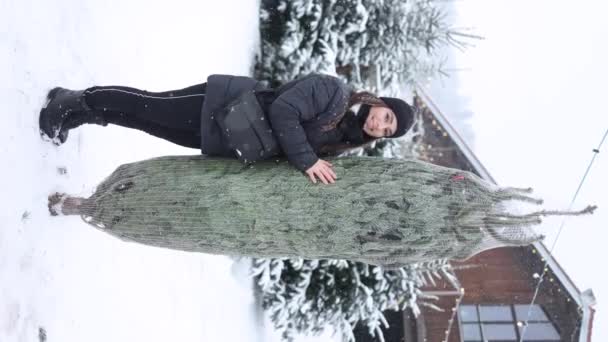 魅力的な若い女性は パックされたクリスマスツリーで立っています 休日を期待して幸せな女性 バーティカルビデオ — ストック動画