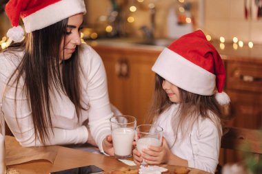 Annesiyle birlikte Noel Baba şapkalı güzel bir kızın portresi mutfak masasının önünde oturmuş süslenmiş peri ışıkları ve taze süt içiyor. Noel havası.
