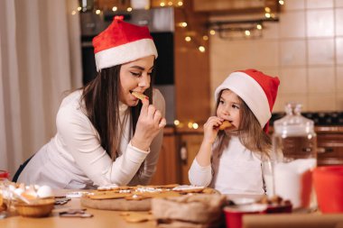 Noel Baba şapkalı sevimli küçük kız ve annesi ev yapımı zencefilli kurabiye yiyip gülüyorlar. Mutlu küçük çocuk Noel için hazırlanıyor. Peri ışıklarının arka planı bokeh.