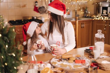 Kadın ve küçük kızı mutfakta lokumla lezzetli kakao içiyorlar. Noel arifesinde ev dekorasyonu. Annem ve Noel Baba şapkalı küçük kız. Peri ışıklarının arka planı. Ağaca sığdır.