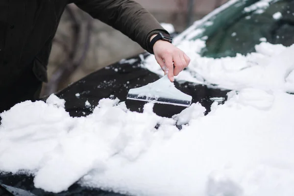 Close up of man is cleaning snowy window on a car with snow scraper. Focus on the scraper. Cold snowy and frosty morning. Black car. High quality photo