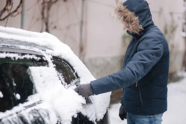 A man is cleaning snowy window on a car with snow scraper. Focus on the scraper. Cold snowy and frosty morning. Black car. High quality photo