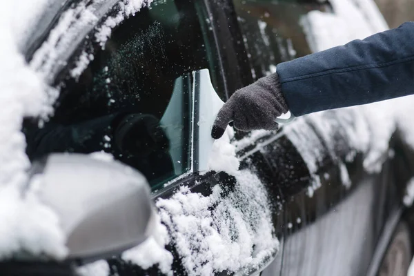 Close up of man is cleaning snowy window on a car with snow scraper. Focus on the scraper. Cold snowy and frosty morning. Black car. High quality photo