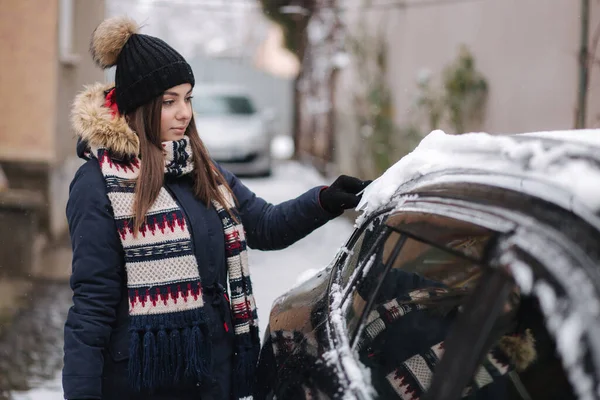 A woman is cleaning snowy window on a car with snow scraper. Pretty woman warmy dressed clean her car outdoors. Cold snowy and frosty morning. Black car. High quality photo