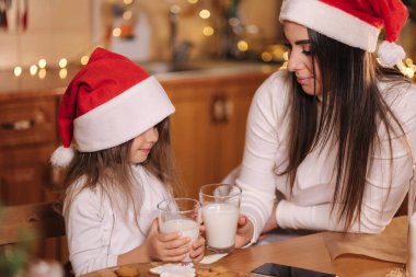 Annesiyle birlikte Noel Baba şapkalı güzel bir kızın portresi mutfak masasının önünde oturmuş süslenmiş peri ışıkları ve taze süt içiyor. Noel havası.