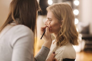 Makyaj sanatçısı güzellik salonunda çalışıyor. Profesyonel makyaj ustası tarafından uygulanan kadın portresi. Güzel makyözler sarı saç modeli için makyaj yapmaya başlar..