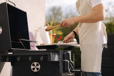 Önlüklü bir adam tavada sebzeleri karıştırmak için tahta kaşık kullanıyor. Izgarada yemek hazırlayın. Yüksek kalite fotoğraf
