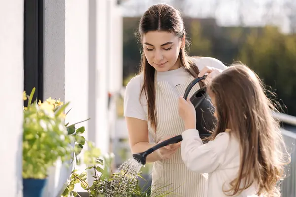 Mom watering plant with her adorable little daughter. Happy family gardening on balcony. High quality photo