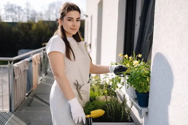 Attractive woman start landing on balcony. Young female in light striped overalls planting. High quality photo