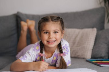 Saç stili örülü sevimli bir kız evdeki kanepede uzanıyor ve annem için resim çiziyor. Yüksek kalite fotoğraf