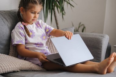 Akıllı küçük kız evdeki kanepede otururken gri dizüstü bilgisayarı açıyor. Yüksek kalite fotoğraf
