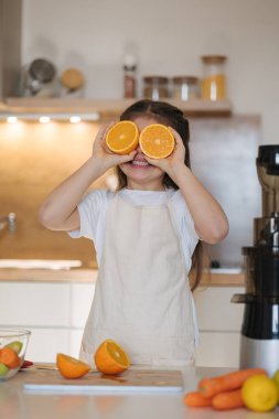 Önlüklü sevimli küçük kızın portresi mutfakta duruyor ve gülümsüyor. Gözlerine portakal yarımları tutuyordu. - Evet. Yüksek kalite fotoğraf