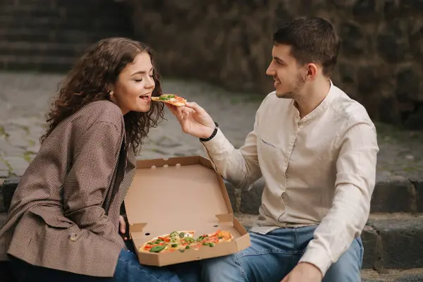 Smiling love couple eating vegan pizza at street on stairs. Handsome man give slice of pizza to woman.