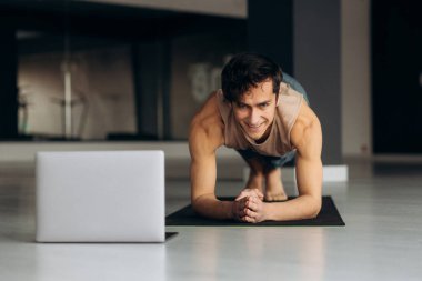 Spor giyim bölümünde vücudunu evde esneten mutlu genç bir adam. Sportif adam dizüstü bilgisayardan özel ders izlerken yoga tahtası yapıyor. Yüksek kalite fotoğraf
