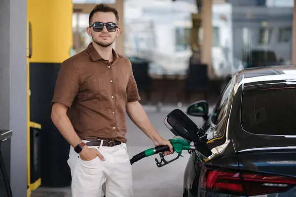 stock image Smiling man refuelling his luxury car at the gas station. Guy looking camera and pouring petrol into tank of modern vehicle on filling station in city. High quality photo