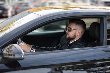Başarı devrede. Deri ceketli, güneş gözlüklü, araba kullanan yakışıklı bir genç. Dışarıdan yan görünüş. Yüksek kalite fotoğraf
