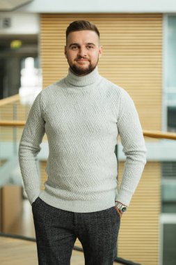 Ofisinin önünde duran şık kazaklı yakışıklı sakallı adamın portresi. Mutlu gülümseyen genç adam. İçeriden bir fotoğraf. Yüksek kalite fotoğraf