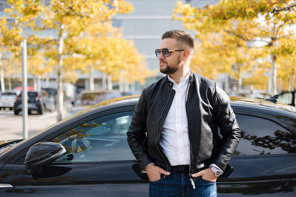 Молодой бизнесмен стоит у своей роскошной машины перед офисом. Стильный бородатый мужчина в черной кожаной куртке. Высокое качество фото