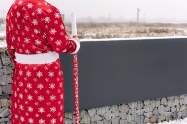 Santa-themed figure in a red snowflake robe holding a staff, standing by a modern stone wall outdoors, festive and unique vibe. High quality photo clipart