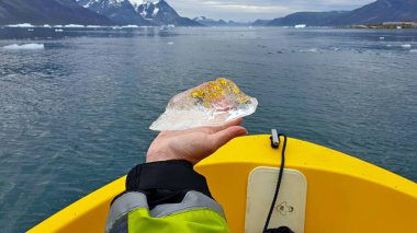 Fiyortta sarı bir botla yüzen dişi bir gezginin eli Grönland 'daki bir buzuldan kopmuş saydam bir milenyum buzunu gösteriyor. Küresel ısınma kavramı