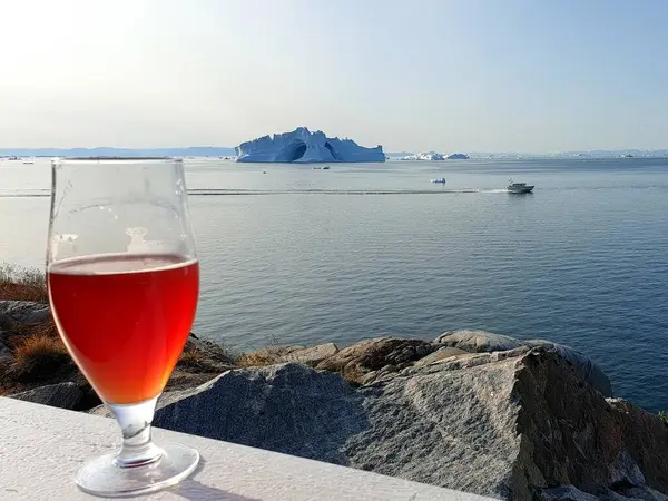 Kutup manzarası, bir bardak birayla denizin arka planına ve Danimarka 'nın batı Grönland şehrinde güneşli bir günde yüzen dev bir buzdağına karşı.