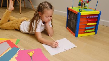 Sevimli küçük liseli kız, evde oyuncaklarla çevrili yaratıcı enerjiyi gösteren mavi kalemle kağıda resim çiziyor. Yüksek kalite 4k görüntü