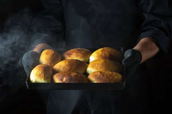 The chef holds a baking sheet with freshly baked buns or pies. Black space for menu or recipe