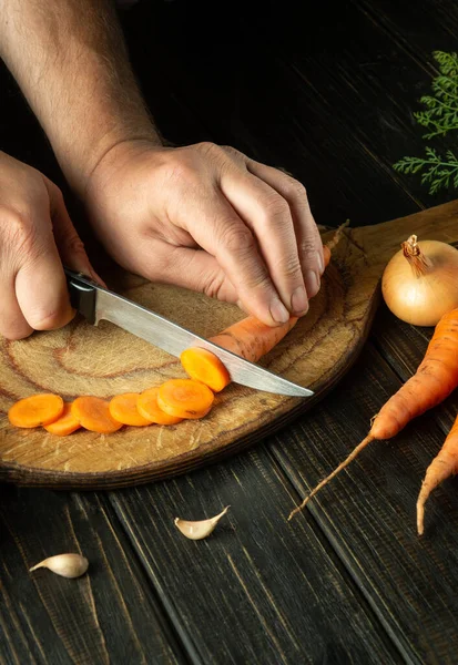 stock image The cook cuts raw carrots with a knife on a wooden cutting board for cooking. Peasant food