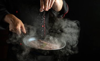 Profesyonel aşçı, sosisli tavaya tuz ekliyor. Siyah arkaplanda otel menüsü veya tarif kavramı