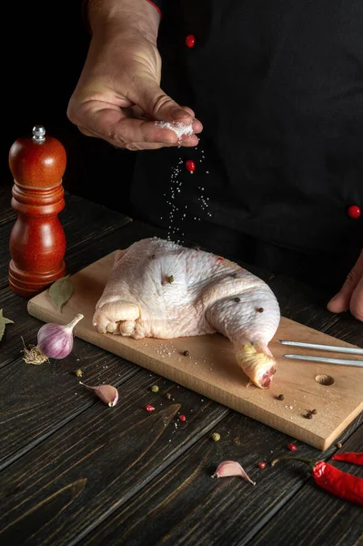 The cook adds salt to the chicken leg. Grill blank with raw chicken leg in the kitchen of a restaurant or hotel on a table with vegetables and spices