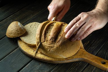 Şef tahtanın üzerinde bir bıçakla buğday ekmeği keser. Mutfak masasında buğday ekmeği mi yoksa sağlıklı beslenme ve geleneksel fırın konsepti mi?.
