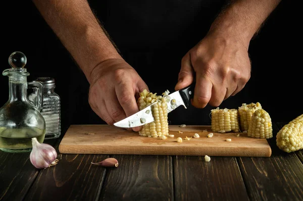 Slicing corn or maize grain by the hands of a chef with a knife on a wooden cutting board. Vegetarian food concept with advertising space