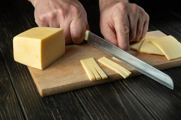 The hands of the cook with a knife cut the cheese into small pieces for tasting on a cutting board. Delicious Milk Cheese Recipe Presentation